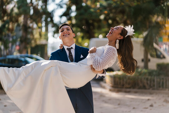 Young groom picking up cheerful bride in park