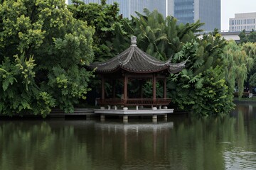 Naklejka premium Serene Pavilion Surrounded by Lush Greenery and Reflective Pond in Urban Park Setting