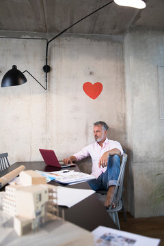 Businessman in a loft using laptop with documents and architectural model on table and heart on the wall