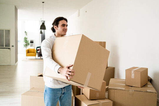Smiling young man carrying cardboard box while moving in new apartment