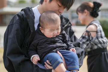 Soft light glows on a tender moment. Man cradles a sleeping baby with care. The child wears a dark jacket and blue pants. Woman stands nearby, smiling gently. The scene feels warm, peaceful