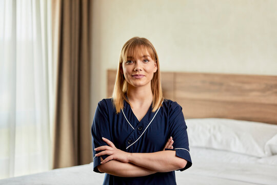 Confident chambermaid with arms crossed standing in bedroom at hotel