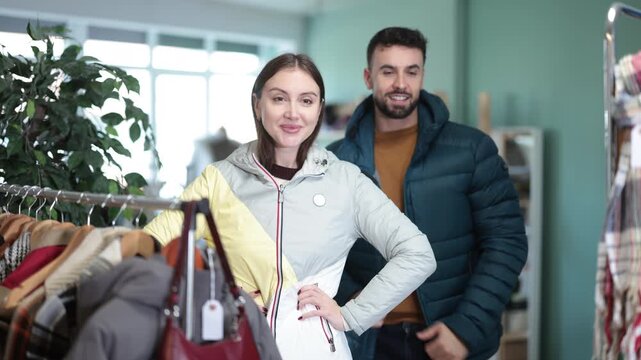Couple stands in a store near a clothes rack and tries on jackets. Buying windbreakers and down jackets in the mall. High quality 4k footage
