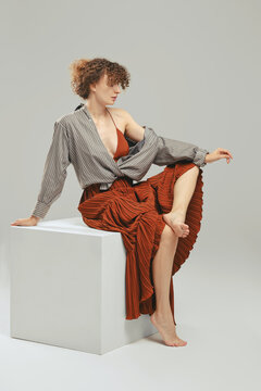 Barefoot woman in sundress and tied up shirt sits on wooden cube in the studio