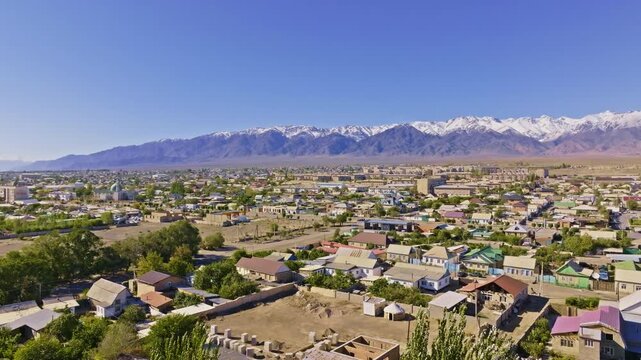Aerial view captures serene Balykchy town surrounded by agricultural land and trees. Snow-capped mountains rise majestically against a clear blue sky.