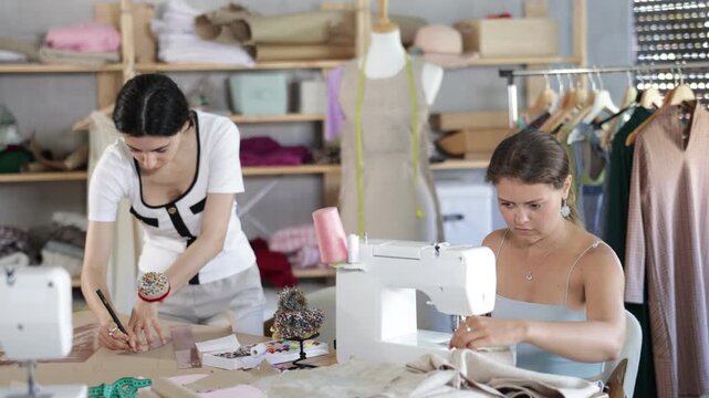 Qualified dressmakers work in a sewing workshop. Fashion designer stands near the table and draws patterns on paper next to the seamstress. High quality 4k footage