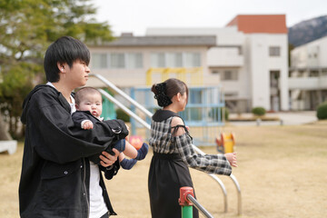 Young man holds baby gently while woman walks nearby. Soft daylight frames peaceful park scene with playground. Family moment suggests calm, everyday connection outdoors. Ideal for parenting, family