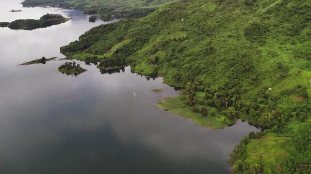 A small unindentifiable boat is moving along the hagged tropical coastline of the Nam Ngam Lake with dense green vegetation in the surrounding.