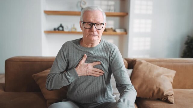 Old man coughing and feeling pain in heart or lung, rubbing chest, portrait. Elderly male person sitting alone in apartment and suffering of heart ache, heart attack or pneumonia, aging of people