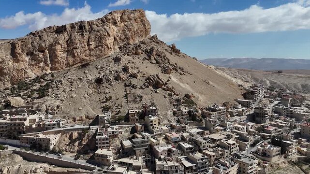Aerial drone shot of the ancient village of Ma'loula, Syria, nestled against the rocky Qalamoun Mountains under a clear blue sky