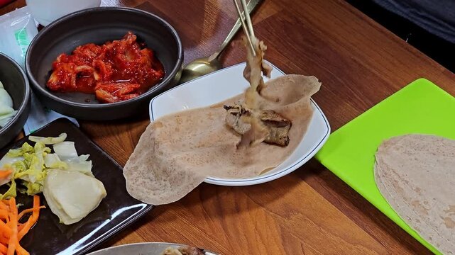 Hands use chopsticks to fill and roll thin buckwheat crepe wrap with boiled pork and vegetables beside kimchi and pickled side dishes in traditional Korean bossam restaurant