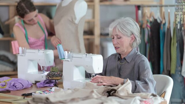 Mature woman is carefully working at a sewing machine and checking the stitches on the fabric against the background of an assistant and a mannequin. Fashion designers working in a workshop. High