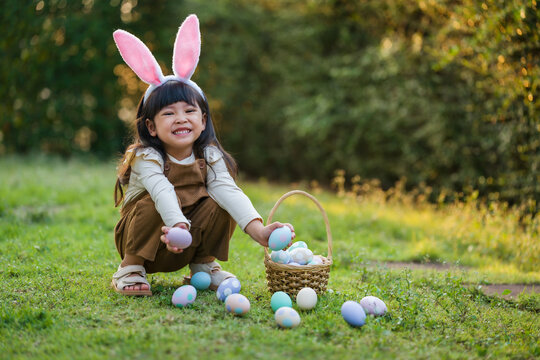 happy child girl wearing bunny ears playing and hunting for Easter eggs into a basket on green grass in garden