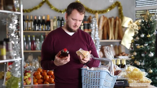 Interested young bearded man in casual maroon sweater with full shopping basket selecting whiskey and cheese in warmly lit grocery store decorated for New Year festivities