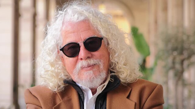 An endearing elderly man with curly white hair and sunglasses exudes confidence while posing with a charming smile, showcasing his unique personality and charisma in a delightful moment captured.