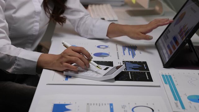 Financial analyst calculating business data and reviewing graphs on a tablet and computer while working overtime in office.