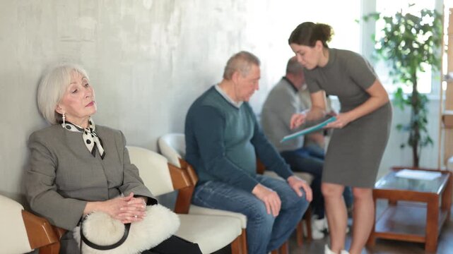 Group of mature people sitting in chairs in queue waiting for appointment, secretary or social worker talking to visitors in background in lobby of public service building 