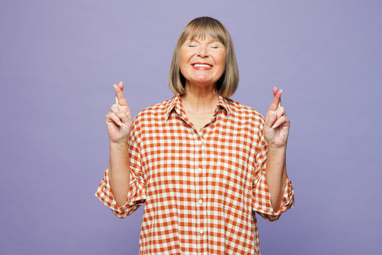 Smiling calm elderly gray-haired woman 50s years old wears casual clothes wait for special moment keep fingers crossed making wish eyes closed isolated on pastel plain pastel light purple background