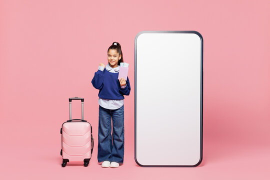 Full body tourist traveler little child kid girl wears blue shirt big huge blank screen area mobile cell phone hold bag passport ticket isolated on pink background. Mother's Day love family concept.