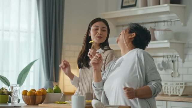 Mother's Day,family bonding,mom day and joyful domestic lifestyle concept.Happy senior Asian mother and adult asia daughter dancing and singing with kitchen utensils while cooking together at home.
