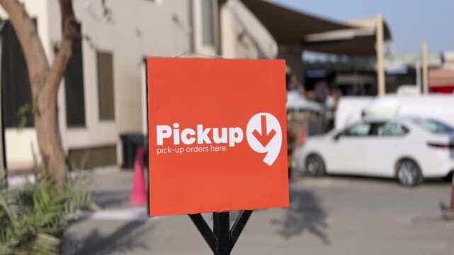 Close-up of an orange pickup orders sign in an outdoor commercial driveway during daylight, with parked car, walkway, and shallow depth of field in the background.