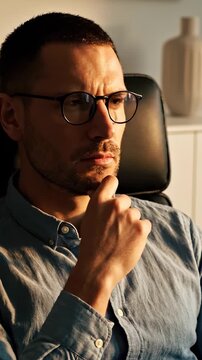 A pensive man thinking hard sits in an office chair. The serious man touches his chin while thinking about a business problem. A pensive look crosses the face of the thinking man. He looks focused.