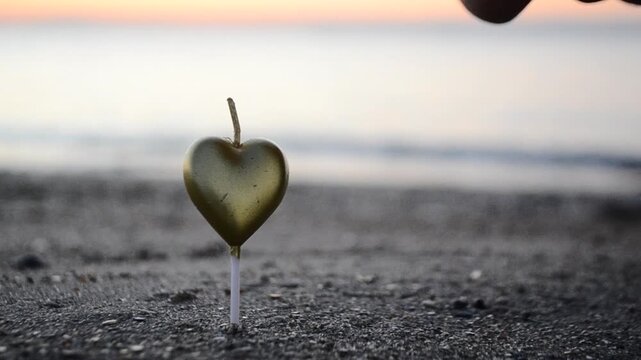 A man lighting a heart-shaped candle in the sand on the beach near the sea at dawn. A man lighting a candle in the shape of heart with lighter standing in the sand on shore near sea at dawn. Sound