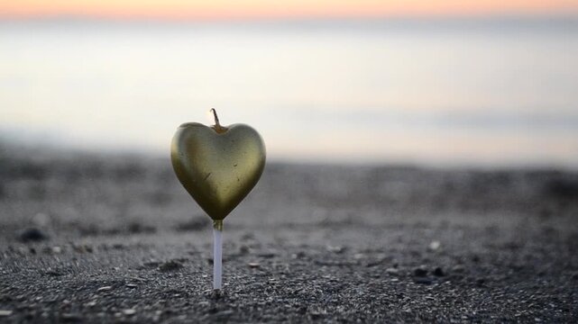 Man lighting heart-shaped candle in sand and covering candle with hand from wind on beach near sea at dawn. Man lighting candle in shape of heart with lighter standing in sand on shore. Sound