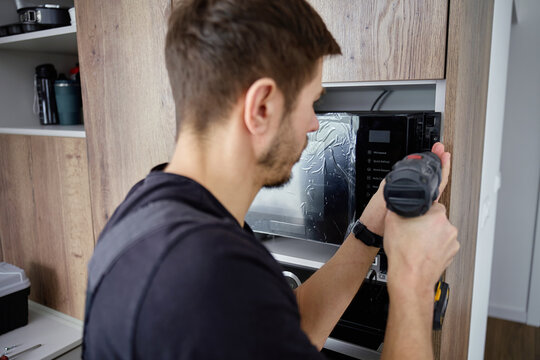 Man using cordless screwdriver to fix built in new microwave oven in modern kitchen cabinet. Concept of home appliance using