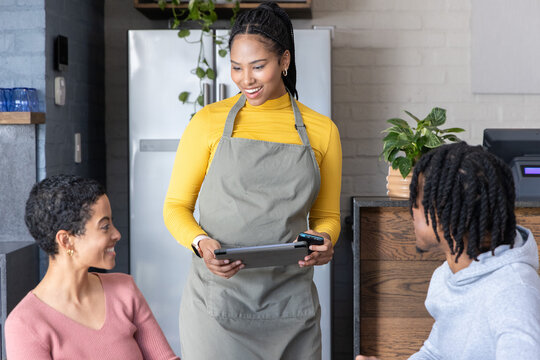 African American female server gray-apron yellow-top serving couple with tablet card-reader at cafe