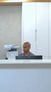 Vertical video: Answering male receptionist using handset at clinic desk, assisting patient via PC