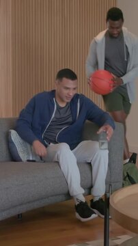 Vertical video: Man in hoodie scrolling phone at home friend entering with basketball causing hug