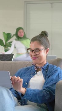 Vertical video: Woman scrolling tablet on studio couch, hijab colleague arranging boards for pitch
