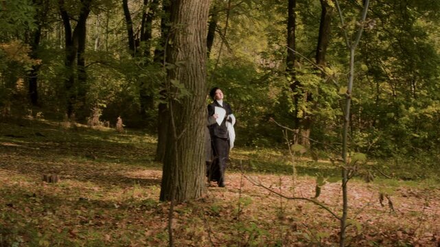During an autumn walk through the forest, a historical couple walks and talks. The woman turns toward the man and walks backward while continuing conversation as the camera follows them in sunlight.