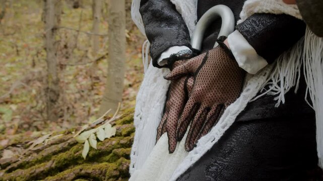 Detailed shot of a woman's hands in black lace gloves over vintage clothing while holding an umbrella in an autumn forest. The scene highlights texture, costume detail and historical style atmosphere.