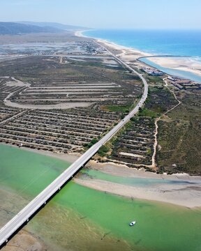 Aerial view of a ribbon of road slicing through vibrant green waters towards the azure sea under a clear sky, Barbate, Andalusia, Spain.