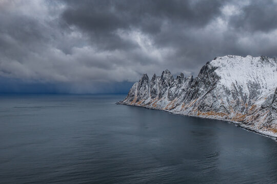 Aerial view of jagged, snow-dusted peaks meeting the somber sea under a brooding sky, Troms, Norway, Senjahopen, Troms, Norway.