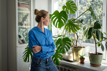 Thoughtful positive middle aged woman florist looking at own indoor garden on windowsill, observing plants with love and care. Mature female plant lover posing surrounded by potted houseplants at home © DimaBerlin