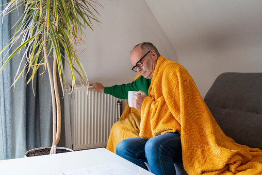 Man with blanket indoors warming up near radiator with hot drink