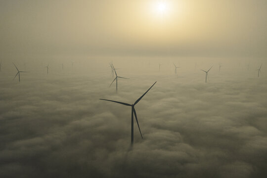 Aerial view of wind turbines piercing through a sea of ethereal fog, with the sun casting a golden glow, Afsluitdijk, Netherlands.