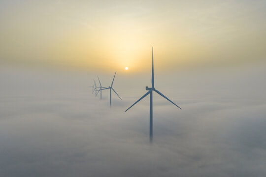 Aerial view of wind turbines piercing through a sea of fluffy clouds under a hazy golden sky, Afsluitdijk, Friesland, Netherlands.