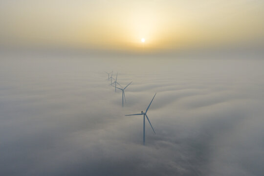 Aerial view of wind turbines piercing through a sea of ethereal fog under a pale, hazy sun, creating a striking contrast of industry and nature, Afsluitdijk, Friesland, Netherlands.