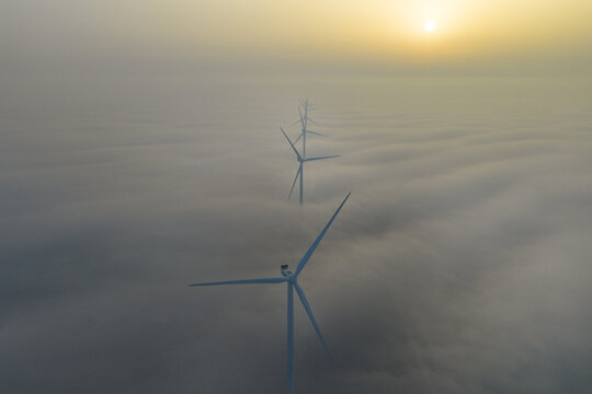 Aerial view of wind turbines rise majestically from a sea of fog, touched by the warm glow of the sunrise, Afsluitdijk, Friesland, Netherlands.