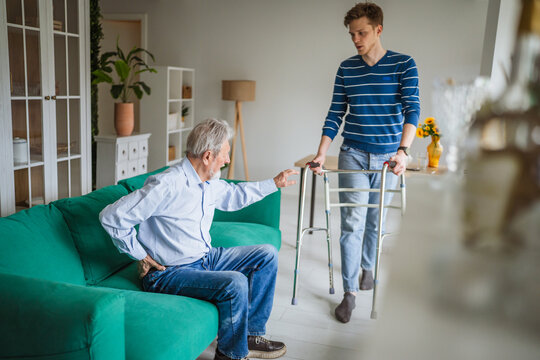 Senior man experiencing back pain at home with caregiver bringing walker