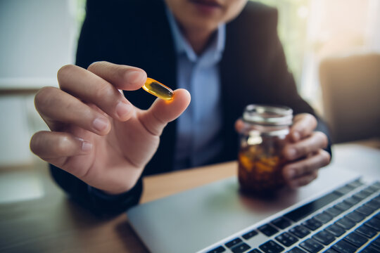Young professional holding a citicoline supplement capsule while working on a laptop at a modern desk. Natural light lifestyle scene illustrating brain health, focus, productivity and workplace 