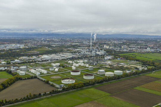 Aerial view of large chemical plant with storage tanks and smoking chimneys, surrounded by green fields in Ludwigshafen, Germany.