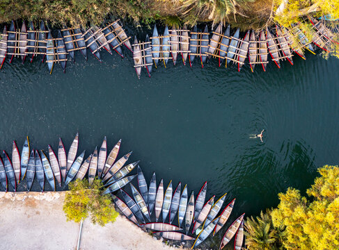 Aerial view of vibrant boats line the tranquil riverbanks near lush greenery, contrasting with the dark water and a lone swimmer, Kineret, Israel.