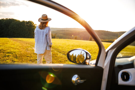 Woman looking at sunset by open car door. Solo traveler relaxing during wanderlust journey. Summer vacation road trip