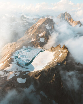 Aerial view of jagged, rocky peaks rise majestically through swirling clouds, capped with stark white snowfields under a soft, ethereal light, Switzerland.