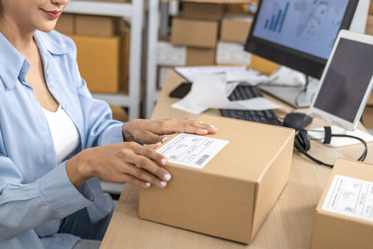 Small business warehouse scene of worker apply shipping label on parcel and taping cardboard box, organizing delivery tasks, order preparation and maintaining efficient packing workflow, multitasking